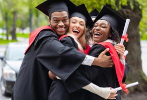 Black guy and two young ladies in graduation costumes posing at camera at university campus, holding diplomas, laughing and hugging, having happy graduation day, closeup portrait photo