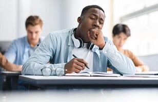 Tired black student yawning sitting at desk in classroom. African American youth exhausted from getting ready for test or writing coursework, feeling sleepy during lecture in highschool. Lack of sleep photo
