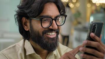 Smiling millennial Indian bearded guy businessman sit in cafe office worker in eyeglasses looking at smartphone screen using mobile phone app scrolling browsing in internet. Arabian man buying online photo