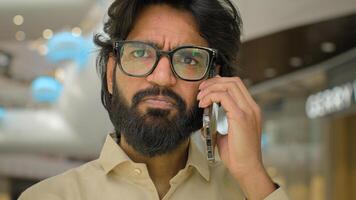 Portrait in office sad Arabian man in glasses talking phone answer business call Close up serious worried Indian male businessman executive CEO speaking distant communication talk mobile conversation photo