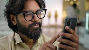 Indian male bearded man in glasses professional hold modern smartphone texting message in office indoors. Arabian businessman using mobile app browsing phone business chatting scrolling web online photo