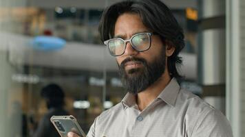 Happy young indian businessman holding smartphone sending message sitting indoors smiling arabian man play mobile game using application on phone reading good news on internet chatting in social media photo