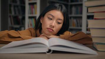 Bored sad tired lazy young korean woman read boring book lying on desk in university library unmotivated female student doing homework preparing for college exam overload with study unloving subject photo