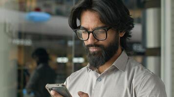 Happy young indian businessman holding smartphone sending message sitting indoors smiling arabian man play mobile game using application on phone reading good news on internet chatting in social media photo