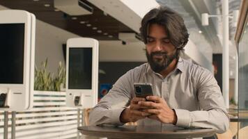 Happy young indian businessman holding smartphone sending message sitting indoors smiling arabian man play mobile game using application on phone reading good news on internet chatting in social media photo