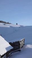 Two rustic cabins sit beneath a thick layer of snow, surrounded by a peaceful winter landscape of rolling hills and evergreen trees under a clear blue sky during midday. Aerial view, vertical footage video