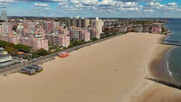 Aerial View of Urban Beach and Boardwalk. Expansive aerial view of a sandy urban beach with a boardwalk, residential buildings, and calm ocean waters under a bright sky. video