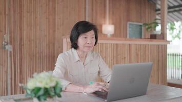 Senior woman using pen to take notes and working on laptop talking on vid call and books on table in bamboo house. Researching. Senior citizen learning to cook online. Self improvement concept. video