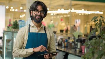 Indian male waiter in cafe working writing client order in digital tablet cafeteria worker Arabian barista man seller in apron noting down menu on wireless computer smiling to camera small business photo
