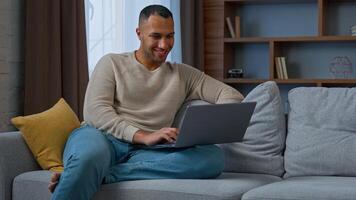 Millennial 30s african american adult man ethnic businessman freelancer sit on couch use pc laptop typing on keyboard distant working chatting online read e-mail e-commerce use home wi-fi networking photo