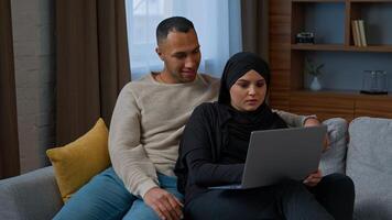 Married couple sit on sofa use laptop device choosing new apartment on internet website multiracial family shocked amazed diverse man woman search information by computer read good news talking indoor photo