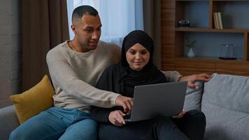 Married couple sit on sofa use laptop device choosing new apartment on internet website multiracial family shocked amazed diverse man woman search information by computer read good news talking indoor photo
