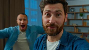 Happy caucasian man blogger vlogger 35s businessman record blog on mobile phone look at webcam camera broadcasting talk indoors on background funny african american friend running fun blogging photo