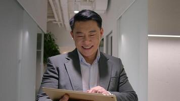 Successful Asian man middle-aged businessman smiling entrepreneur walking at office hallway go to business meeting use digital tablet portable computer device browsing website social networks chatting photo