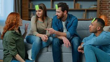 Group of multiethnic friends play charades game at home happy laughing diverse ethnic men and women guessing guess personage playing with sticky notes on head talk ask questions laugh entertainment photo