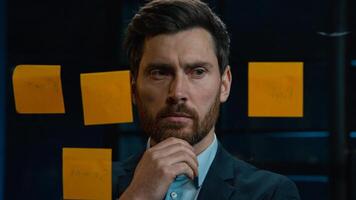 Thoughtful Caucasian businessman employee middle-aged man analyze information on stickers read sticky notes on glass wall standing in office planning project manage tasks startup ideas organizes work photo