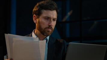 Caucasian 40s businessman employee auditor middle aged man sit in evening night office read business documents papers legal contracts pensive employee think check data in computer search information photo