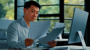 Professional asian businessman korean japanese 40s man lawyer office worker reading financial documents invoice contract budget documentation searching business data at computer paying bills online photo