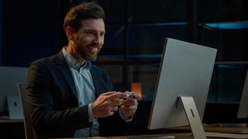 Adult businessman employee working in evening office happy male gamer Caucasian man take break playing console game on computer with gaming controller using joystick gamepad play virtual match photo
