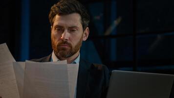 Caucasian 40s businessman employee auditor middle aged man sit in evening night office read business documents papers legal contracts pensive employee think check data in computer search information photo
