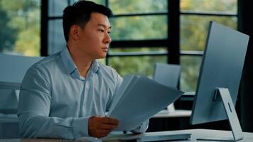 Professional asian businessman korean japanese 40s man lawyer office worker reading financial documents invoice contract budget documentation searching business data at computer paying bills online photo