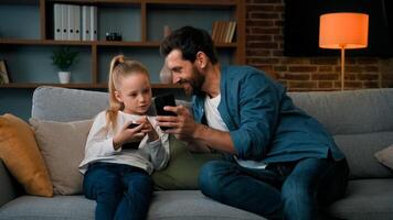 Caucasian man and little kid girl sit together at home hold modern phone browsing media content play games online adult dad teach child daughter use mobile apps showing funny on wireless device photo