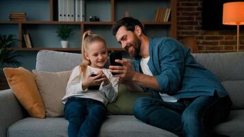 Caucasian man and little kid girl sit together at home hold modern phone browsing media content play games online adult dad teach child daughter use mobile apps showing funny on wireless device photo