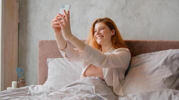 Happy Caucasian woman in bed talking call chat with mobile phone app thumb up waving hello goodbye smiling online conference carefree girl blogger vlogger record stream vlog at morning bedroom photo