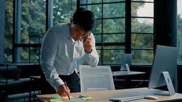 Busy asian businessman korean man working stand at table in modern office talk on phone give professional consultation by mobile call write tasks on sticky notes project paperwork multitasking concept photo