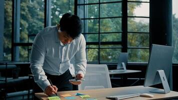 Senior asian ethnic man entrepreneur businessman stand at table in modern office use mobile phone write tasks on sticky notes organize paperwork project business plan multitasking work brainstorming photo