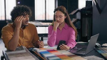 Two designers male female colleagues in modern design studio looking at fabric samples discussing color textile for interior angry Arabian Indian man talk with Caucasian woman problem misunderstanding photo