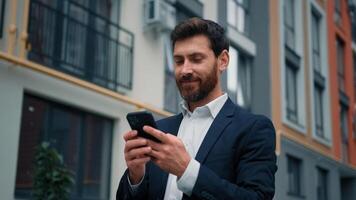 Attractive caucasian businessman stand in city texting messages on mobile phone positive smiling 30s freelancer owner read internet digital news good notification browsing media content online working photo