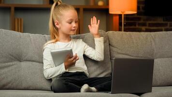 Blonde little smart schoolgirl pupil listen educational lesson on laptop at home online class meeting in chat during pandemic write homework tasks at copybook raise hand ready to answer question photo