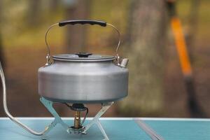 A silver kettle sits atop a portable stove, ready for use in a tranquil forest setting surrounded by nature photo