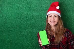 beautiful girl in a New Year's cap on a green background shows a phone screen with a chromakey background. Christmas. New Year photo