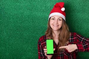 beautiful girl in a New Year's cap on a green background shows a phone screen with a chromakey background. Christmas. New Year. photo