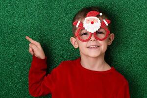 a cute boy in New Year's souvenir glasses with Santa Claus on a green background points to the background. New Year photo