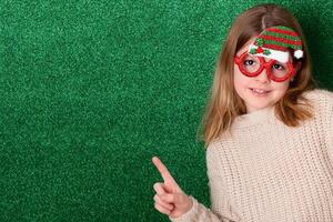 a cute girl in New Year's souvenir glasses with a New Year's cap on a green background points to the background. New Year photo