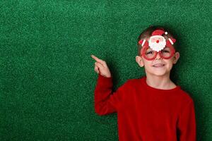 a cute boy in New Year's souvenir glasses with Santa Claus on a green background points to the background. New Year photo