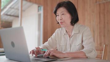 Senior woman using laptop talking on call and holding pen and book on table in bamboo house.Self development concept. video