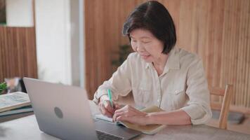Close up of senior woman face using pen to take notes and using laptop to talk on call on table in bamboo house. Researching, senior citizen learning to cook online. Self development concept. video