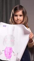 vertical portrait of a Little girl is holding a piece of paper with a rabbit on it. Smiling brightly, girl showcases her artistic talent and creativity in a warm, inviting space video