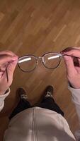 vertical pov First person view Man putting on glasses. concept of deteriorating vision. Man casually holds stylish eyeglasses over a clean wooden floor, examining frames and lenses video
