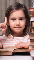 vertical little girl had a good idea. The girl shows a gesture of raising her index finger up. a portrait of a little girl surrounded by stacks of books in a library, in an apartment video