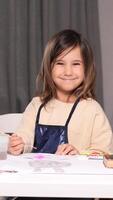 vertical portrait of a little girl drawing with paints and a paintbrush. The girl looks at the camera and smiles. Looking at the camera video