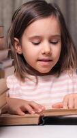 vertical little girl is sitting and reading a book with two tall piles of books on each side of her. She is smiling at the book as she reads. close-up of a little girl reads a book out loud video