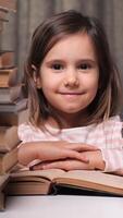 vertical portrait of a little girl surrounded by stacks of books in a library, in an apartment. The girl looks at the camera and smiles, an open book lies in front of her. The concept of learning read video