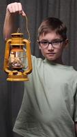 vertical Boy wearing eyeglasses and casual clothes holding a yellow kerosene lamp illuminating a dark room with gray curtains in the background video
