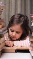 vertical little girl reading a book behind a table full of books. Stacks of books surround the little girl. Child learning to read, Girl runs her fingers along the lines of a paper book video