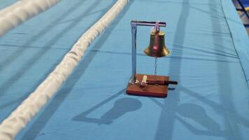 Boxing bell sitting on blue canvas beside ring ropes, casting shadows under bright lighting, ready to signal start or end of round video